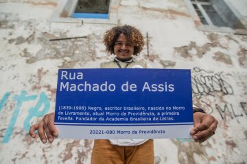 Cosme com uma placa de rua em homenagem a Machado de Assis